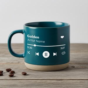 May include: A dark teal ceramic mug featuring a white graphic print of a music player interface. The text reads Golden and Artist Name, with a progress bar, heart icon, and playback controls including shuffle, skip, pause, and repeat symbols.