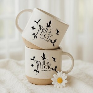 May include: Two stacked ceramic mugs with a speckled white finish and a tan base. Each mug features black bat illustrations and the text "Well the FUCK aware". A white daisy with a yellow center sits near the bottom mug.