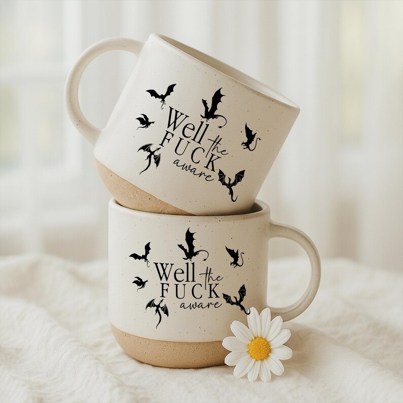 May include: Two stacked ceramic mugs with a speckled white finish and a tan base. Each mug features black bat illustrations and the text "Well the FUCK aware". A white daisy with a yellow center sits near the bottom mug.
