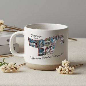 Op de afbeelding: Een witte keramische mok met een gespikkelde afwerking en een beige basis. De voorzijde bevat de tekst Greetings from WIDOW'S BAY ...The next Martha's Vineyard! met kleurrijke grafische illustraties van mensen en gebouwen.