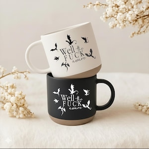 May include: Two ceramic mugs stacked on top of each other. The top mug is white with a brown base, the bottom mug is black with a brown base. Both mugs have a dragon design and the text "Well the FUCK aware".