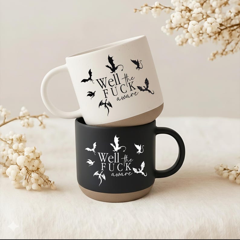 May include: Two ceramic mugs stacked on top of each other. The top mug is white with a brown base, the bottom mug is black with a brown base. Both mugs have a dragon design and the text "Well the FUCK aware".