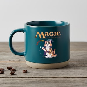 Può includere: Tazza in ceramica color verde acqua con base beige, con un design a fumetto di un gatto mago e la scritta "Magic The Cattering". La tazza è cilindrica con un grande manico. Chicchi di caffè sparsi.