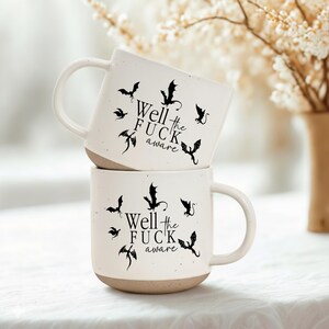 May include: Two white ceramic mugs stacked on top of each other. Each mug features black bat illustrations and the phrase "Well the FUCK aware". The bottom of each mug is a light brown color, and the mugs have a speckled texture.