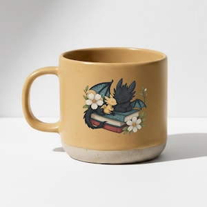 May include: A mustard yellow ceramic mug with a speckled finish and a base in a lighter, matte beige tone. The front features a graphic illustration of a small yellow dragon and a larger black dragon resting on a stack of books with white flowers.