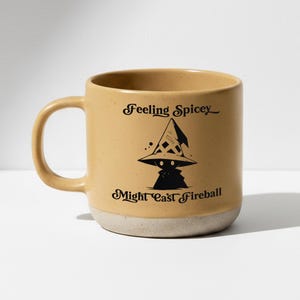 Könnte beinhalten: Ein beigefarbener Keramikbecher mit gesprenkelter Oberfläche und hellgrauem Boden. Der Becher zeigt eine schwarze Grafik eines Zauberhuts und einer kleinen Figur, mit dem Text "Feeling Spicey" darüber und "MIGHT CAST FIREBALL" darunter in schwarzer Schrift.