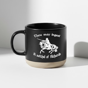 May include: A black ceramic mug with a beige base featuring a white line-art illustration of a knight on horseback. The text "Thou may ingest A satchel of Richards" is printed in a white gothic-style font above and below the illustration.