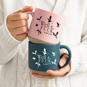 May include: Two ceramic mugs, one pink and one teal, stacked together. The pink mug has black dragon illustrations and the text "Well the FUCK aware." The teal mug has white dragon illustrations and the same text. Both mugs have a tan base.