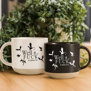 May include: Two ceramic mugs, one white and one black, with a beige base. Each mug features the phrase "Well the FUCK aware" in a stylized font, surrounded by bat silhouettes. The mugs are set against a backdrop of green foliage.