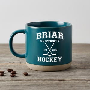 Puede incluir: Taza de cerámica color turquesa con base beige. La taza presenta el texto "Briar University Hockey" con palos de hockey cruzados. La taza está sobre una superficie de madera con granos de café.