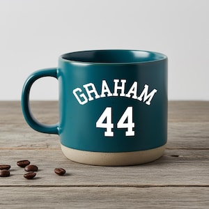 Puede incluir: Taza de cerámica color turquesa con el nombre "GRAHAM" y el número "44" en letras blancas. La parte inferior de la taza es de color beige claro. Varios granos de café están esparcidos sobre una superficie de madera junto a la taza.