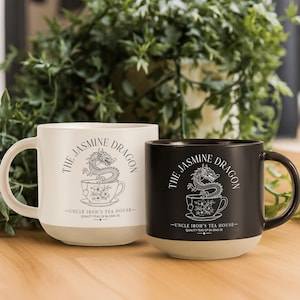 May include: A white ceramic mug and a black ceramic mug, each featuring a graphic of a dragon in a teacup with the text The Jasmine Dragon, Uncle Iroh's Tea House, Quality Teas of Ba Sing Se. Both mugs have a smooth, unglazed beige base.