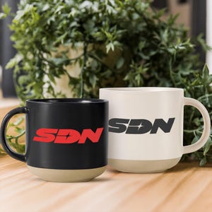 May include: A black ceramic mug with a red SDN logo and a white ceramic mug with a dark grey SDN logo. Both mugs feature a rounded handle and a contrasting unglazed beige base. The logo on each mug includes a small four-pointed star icon.