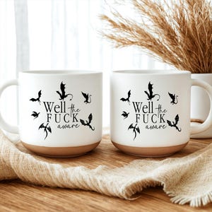 May include: Two white ceramic mugs with a brown base. Each mug features the phrase "Well the FUCK aware" in black text, surrounded by black dragon illustrations. The mugs are on a wooden surface.