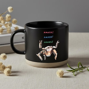 May include: A black ceramic mug with a beige base featuring a graphic of a multi-limbed creature in a dynamic pose. Above the creature, the word "AMAZE!" is printed three times in stacked rows, using red, blue, and green text respectively.