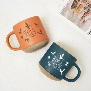 May include: Two ceramic mugs, one orange and one teal, with a tan base and handle. Each mug features a dragon graphic and the text "Well the FUCK aware". The mugs are on a white, fluffy surface.