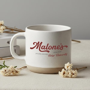 Puede incluir: Taza de cerámica blanca con acabado moteado y base beige. La taza presenta el texto "Malone's" en escritura burdeos, junto con "proudly serving Briar University since 2015."