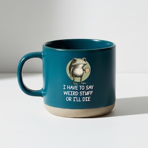 Pode incluir: Uma caneca de cerâmica azul-petróleo com base bege, apresentando um gráfico de um sapo segurando uma pequena xícara dentro de um círculo verde-claro. Abaixo do sapo, o texto diz: I HAVE TO SAY WEIRD STUFF OR I'LL DIE. A caneca tem uma alça curva.