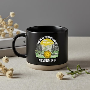 Może przedstawiać: Czarny ceramiczny kubek z beżową podstawą, ozdobiony grafiką żółtej dwudziestościennej kości do gry na tle górskiego krajobrazu. Napis "NOT ALL THOSE WHO WANDER ARE LOS... NEVERMIND" widnieje białymi i żółtymi literami na przedniej stronie.