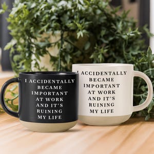 Könnte beinhalten: Zwei Keramikbecher, einer schwarz und einer weiß, jeweils mit dem schwarzen oder weißen Textaufdruck: I ACCIDENTALLY BECAME IMPORTANT AT WORK AND IT'S RUINING MY LIFE. Beide Becher haben einen glatten oberen Teil und einen hellbraunen Boden.