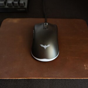 May include: A black computer mouse with a white accent and a bat logo rests on a brown leather mousepad. The mousepad has rounded corners and a small embossed logo in the corner. A keyboard is visible in the background.