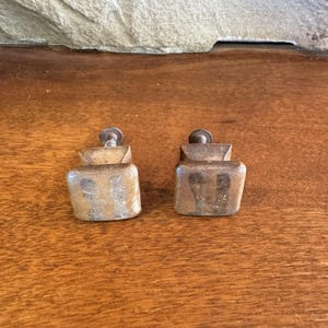 May include: Two vintage wooden cabinet knobs with a square base and a metal screw. The knobs have a weathered, aged appearance, with visible wood grain and a warm, brown color. They are likely for a drawer or cabinet.