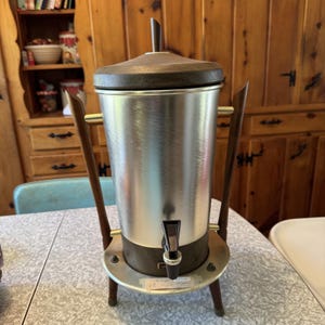 May include: A vintage stainless steel coffee urn with a dark brown lid and base. The urn has a spigot and sits on a wooden stand with three legs. The urn is on a patterned table.