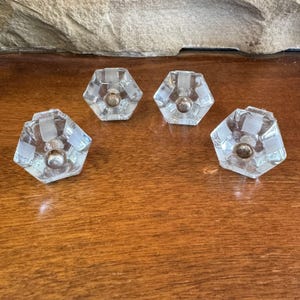 May include: Four clear glass cabinet knobs with a hexagonal shape and a silver-colored center. The knobs have a textured, striped design on the sides. They are arranged on a wooden surface.