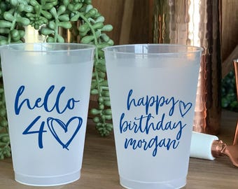 Può includere: Due bicchieri di plastica smerigliata con testo blu. Uno dice "hello 40" con un cuore, l'altro dice "happy birthday morgan". Sullo sfondo uno shaker per cocktail, un jigger e un apribottiglie in rame.