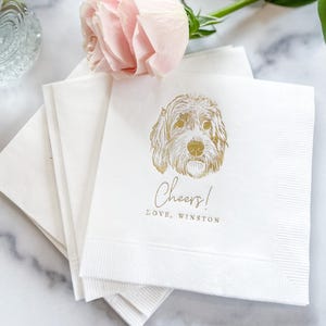 May include: White cocktail napkins with a gold illustration of a dog's face, the word "Cheers!" and the text "LOVE, WINSTON". A pink rose and greenery are in the background. Clear glass drinkware is also visible.