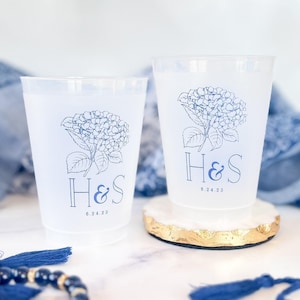 May include: Two frosted plastic cups with a blue hydrangea design, the initials "H&S", and the date "6.24.23". One cup rests on a white and gold coaster. A blue beaded bracelet and fabric are in the background.