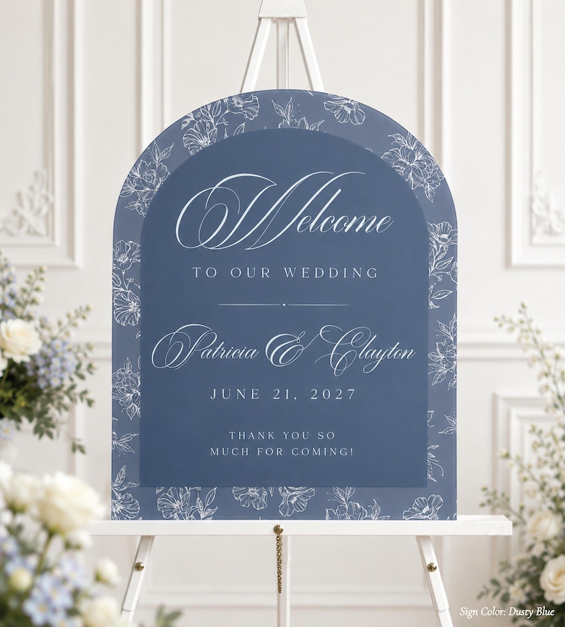May include: A dusty blue wedding welcome sign with floral accents. The sign reads "Welcome to our Wedding" and includes the names Patricia & Clayton, and the date June 21, 2027. The sign also says "Thank you so much for coming!"