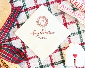 May include: A square white napkin with a red wreath design and the words "Merry Christmas" and "The Fife's" printed in red. The napkin is on a red, green, and white plaid tablecloth, with a wooden spoon and candy canes.