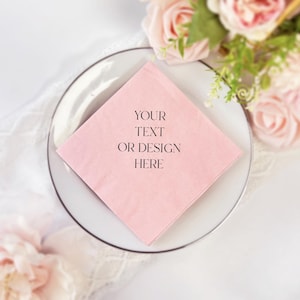 May include: A light pink square napkin with the text "YOUR TEXT OR DESIGN HERE" printed in black, placed on a white plate with a silver rim. The background includes soft pink roses and greenery, suggesting a wedding or event theme.