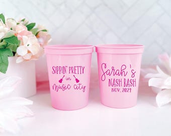 May include: Two pink plastic cups with text. One cup reads "Sippin' Pretty in Music City" with guitar graphics. The other cup says "Sarah's Nash Bash Nov. 2021". The cups are on a pink surface with flowers in the background.