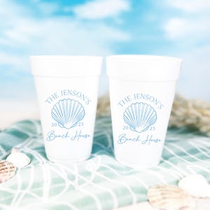 May include: Two white foam cups with a blue seashell design and the text "The Jenson's 2025 Beach House". The cups are on a blue and white striped towel with seashells scattered around.