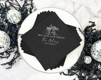 May include: A stack of black square napkins on a white plate. The napkins have the text "NEW YEAR, SAME FRIENDS, The Ashers, NYE PARTY" in a silver font, with a graphic of two champagne glasses clinking. Decorative disco balls and black paper filler surround the plate.