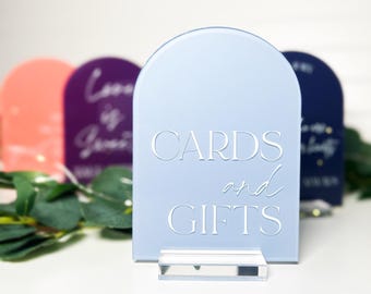 May include: A light blue arched acrylic sign with the white text CARDS and GIFTS, mounted on a clear rectangular base. The sign features a smooth, matte finish and is positioned in front of other blurred, colorful arched signs.