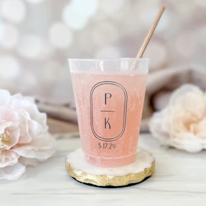 May include: A clear plastic cup filled with a pink beverage and a wooden straw. The cup has a black oval design with the letters "P" and "K" and the date "5.17.24". The cup sits on a white and gold coaster.