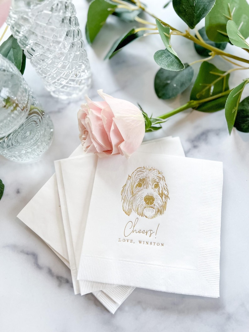 May include: White cocktail napkins with a gold illustration of a dog's face, the word "Cheers!" and the text "LOVE, WINSTON". A pink rose and greenery are in the background. Clear glass drinkware is also visible.