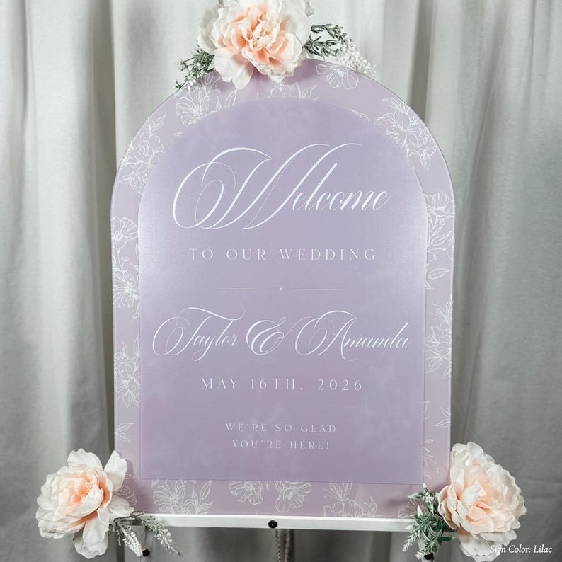 May include: A lilac-colored wedding welcome sign with floral accents. The sign features the words "Welcome to our wedding", the names "Taylor & Amanda", and the date "May 16th, 2026". The sign is decorated with flowers.