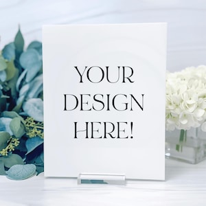 May include: White sign with a black stand, featuring the text "YOUR DESIGN HERE!" on a white background. The sign is sitting on a white surface with greenery and white flowers in the background.