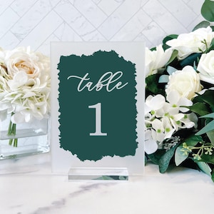 May include: A white acrylic table number with a dark green painted background. The number "1" and the word "table" are written in white on the background. The table number is on a clear acrylic stand.