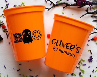 Può includere: Due bicchieri di plastica arancioni. Una tazza ha la parola "BOO" con una grafica a fantasma. L'altra tazza ha il testo "OLIVER'S 1ST BIRTHDAY" in nero. Articoli per feste di Halloween.