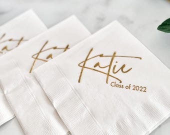 以下が含まれることがあります： 金色の文字が印刷された白い紙ナプキン。名前「Katie」と「Class of 2022」がパーソナライズされています。折りたたまれたナプキンが積み重ねられており、卒業のお祝いに最適です。