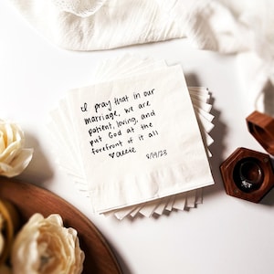 May include: A stack of white paper napkins with handwritten text: "I pray that in our marriage, we are patient, loving, and put God at the forefront of it all." A wooden ring box and a ring are visible.