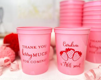Puede incluir: Vasos de plástico rosa con texto rojo y gráficos de fresas. Una taza dice "Thank you berry much for coming!" y la otra dice "Caroline is TWO sweet." Hay pilas de vasos rosas en el fondo.