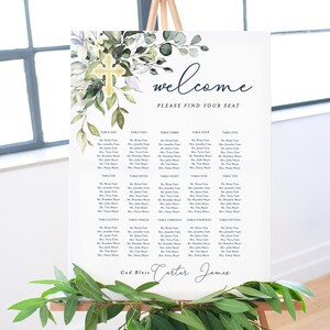 Puede incluir: Un diagrama de asientos de boda blanco con una cruz dorada y follaje verde. El diagrama enumera las mesas del uno al quince con los nombres de los invitados. El texto en la parte inferior dice "God Bless Carter James".