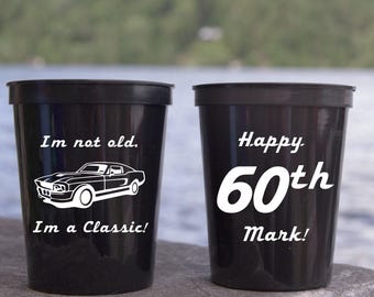 May include: Two black plastic cups with white text. One cup says "I'm not old, I'm a Classic!" with a car graphic. The other says "Happy 60th Mark!" These cups are suitable for a birthday celebration.