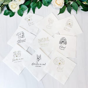 May include: A collection of white paper napkins with various dog illustrations in black and gold ink. Each napkin features a different dog breed and a personalized message such as "Cheers!" or "Drinks on me!" with a name.